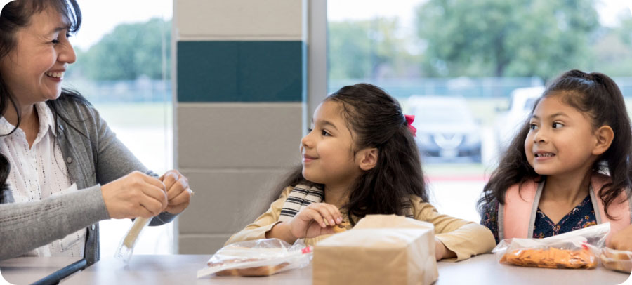 Teacher helping a kid open a string cheese while another child looks on.