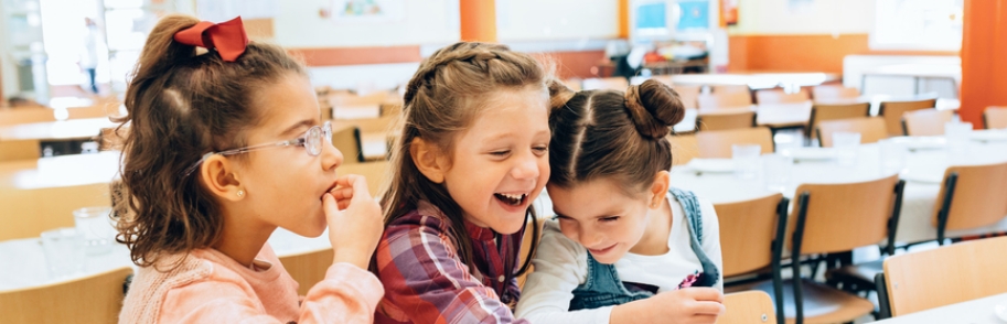 Three children sit together at a cafeteria table in a brightly lit school room
