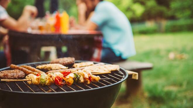 Food on a barbecue with people at a picnic table in the background